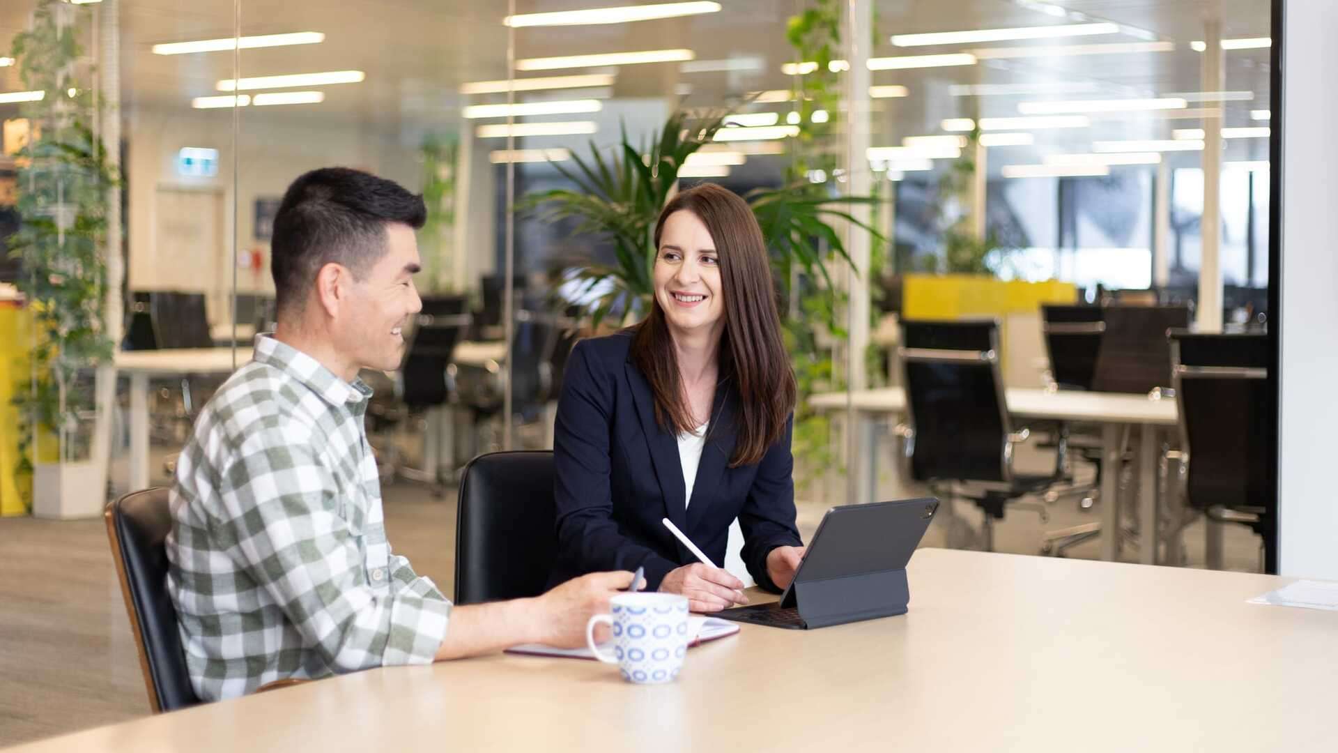 Two students sitting at a table talking while one works on a tablet.