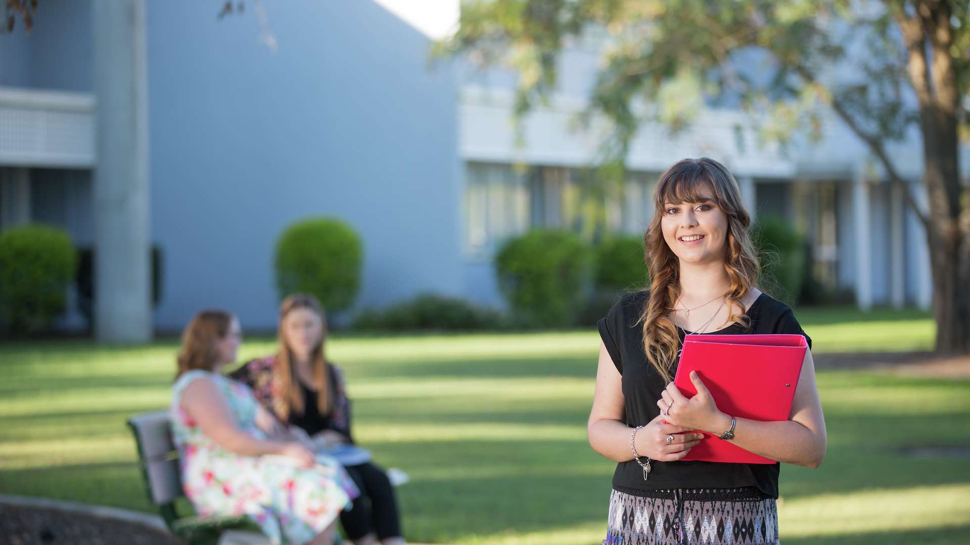 Student holding folder and smiling with two students sitting in background on Bundaberg campus