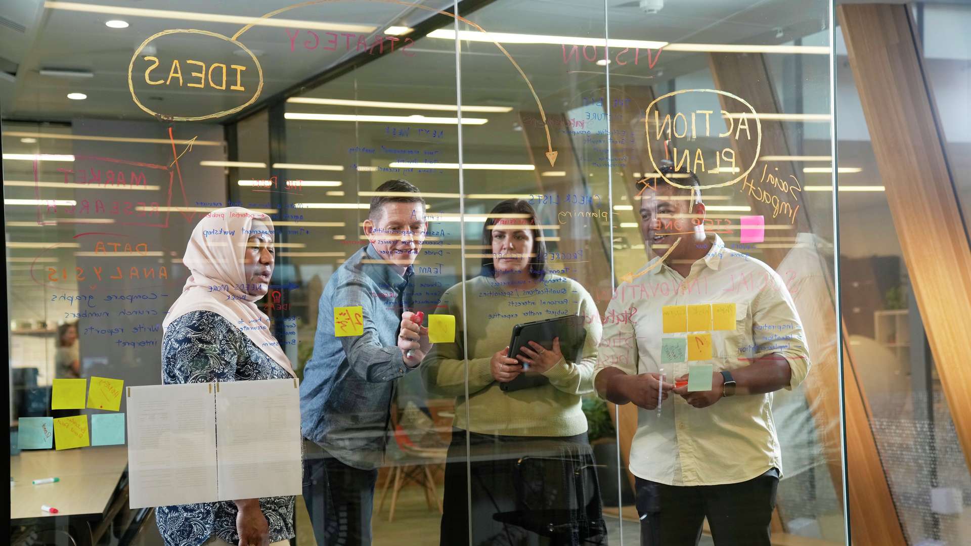 A group of four students writing on glass discussing plans and putting sticky notes up.