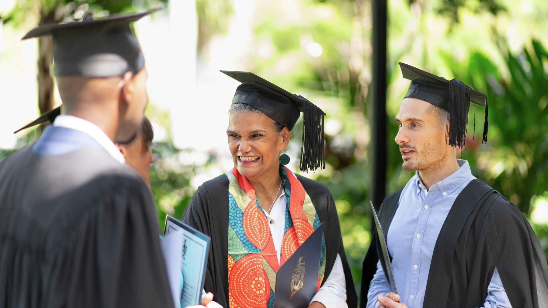 Four CQU students wearing graduation regalia and hold their testamurs as they smile and chat.