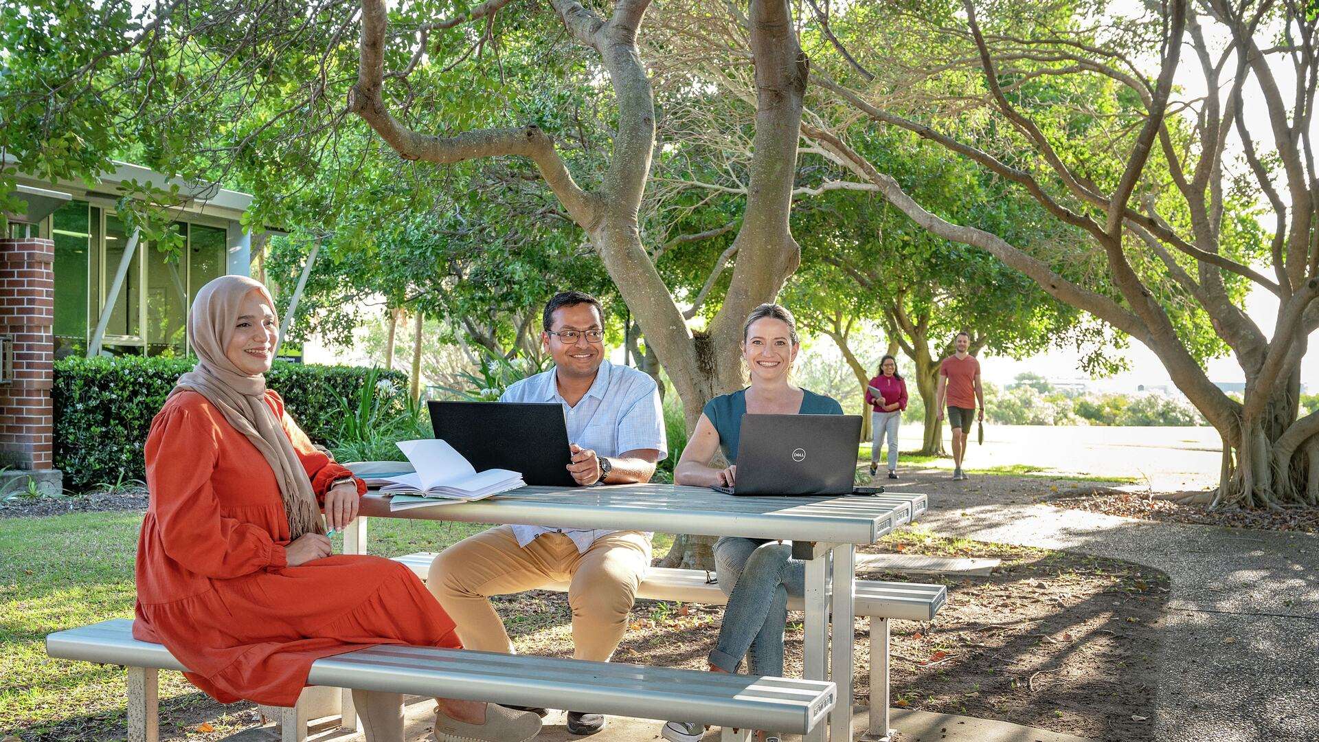 Three students sitting at table on Gladstone campus studying and two students walking down path in background