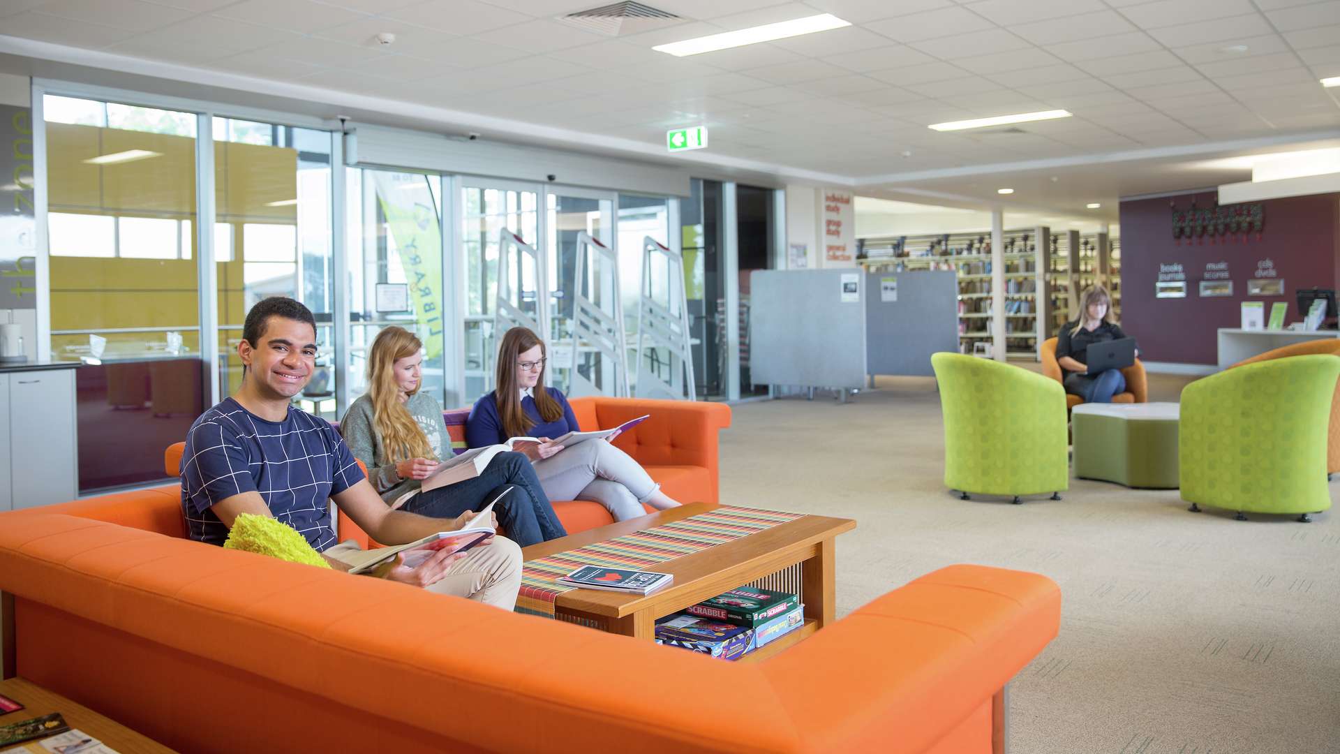 A student reading a magazine sits on a couch and smiles while other students read books nearby.
