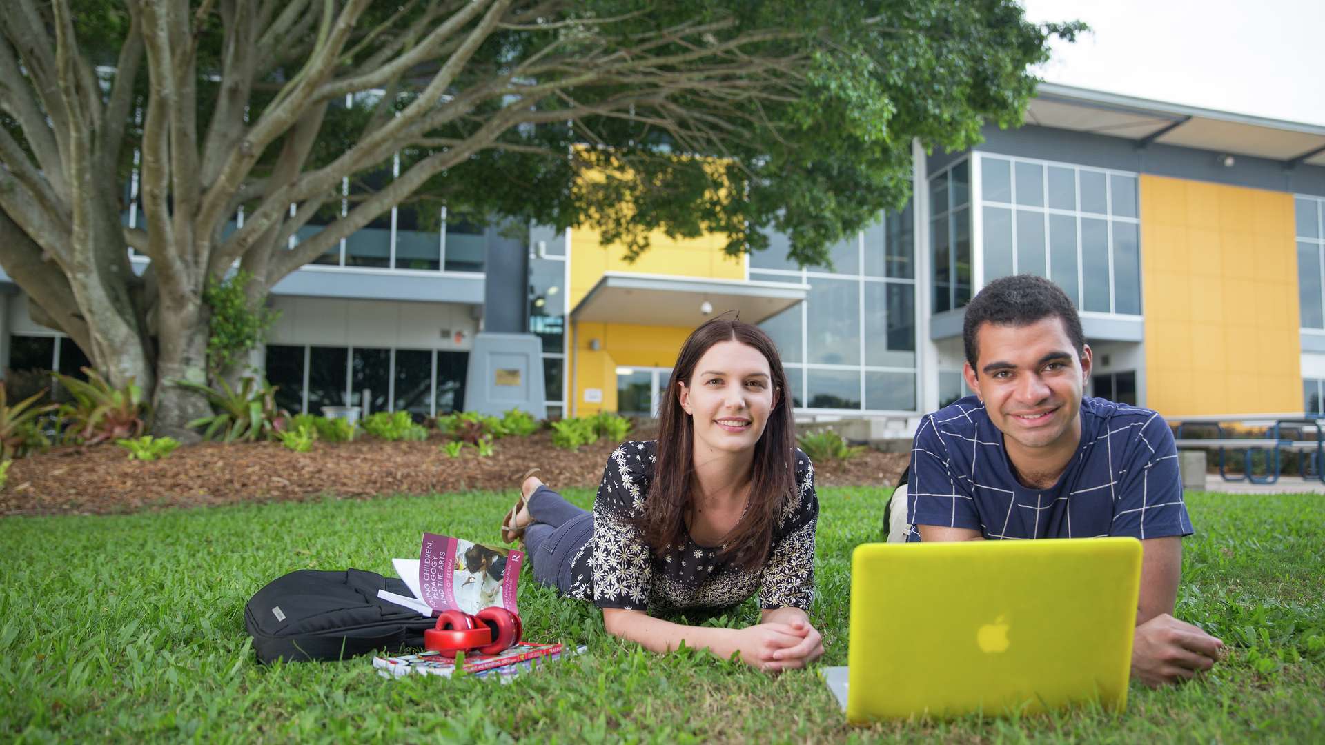 Two students lying on grass at Mackay campus with yellow laptop.