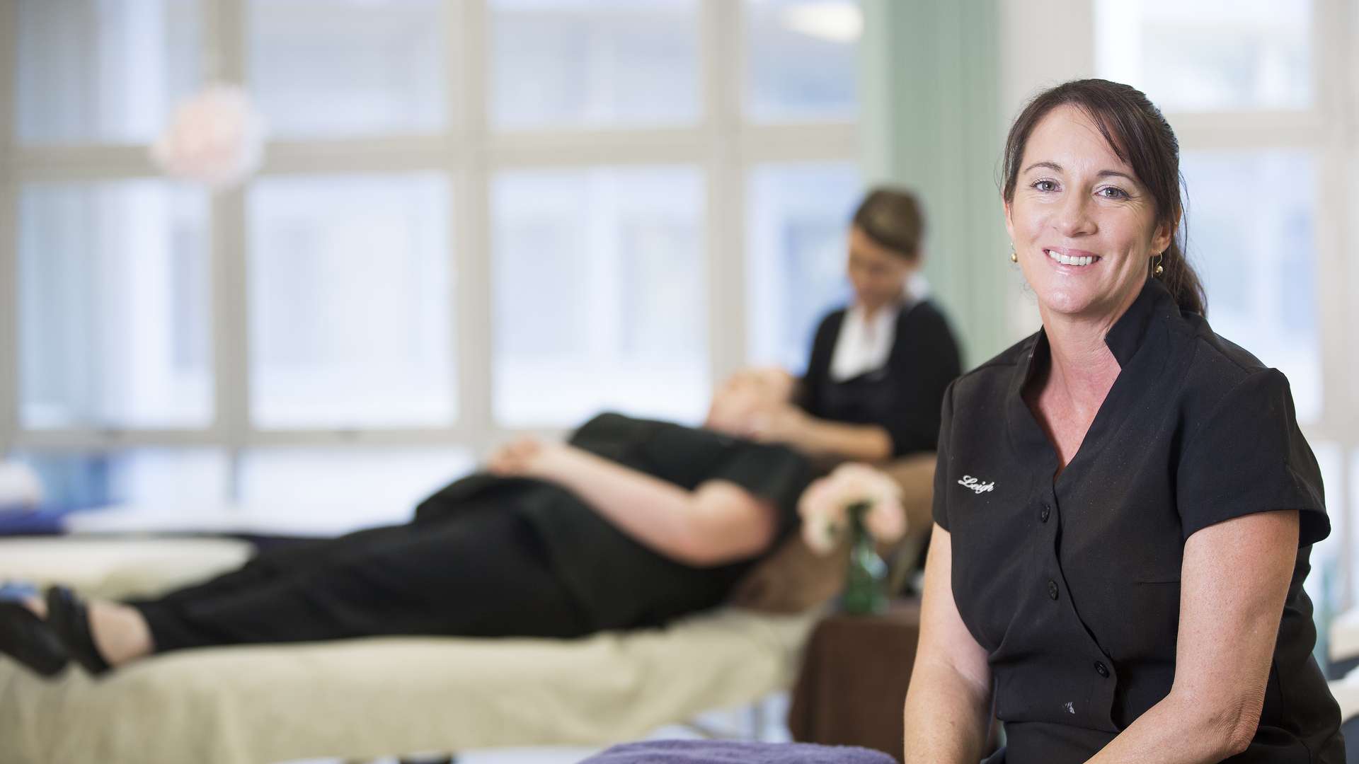 Beauty student smiling at the camera as another student gives a client a facial in the background