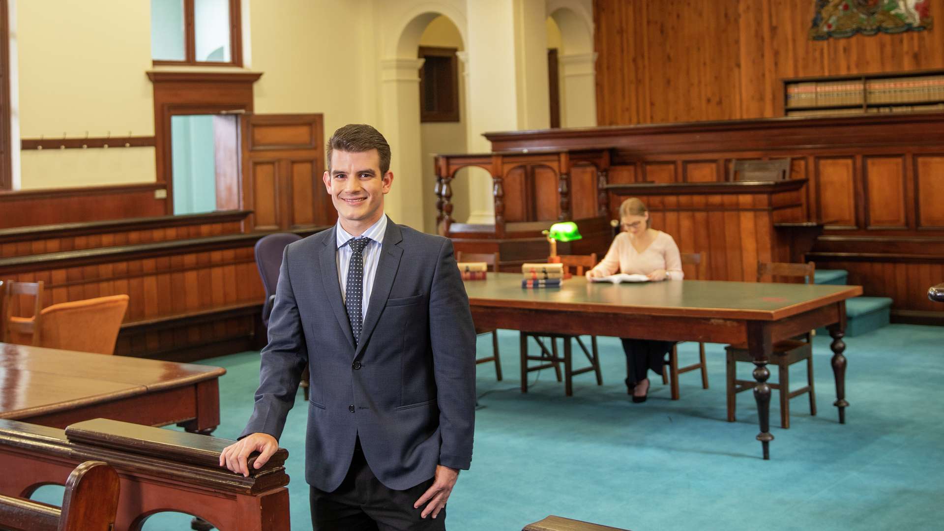 Lawyer stands in courtroom with law student in the background studying.