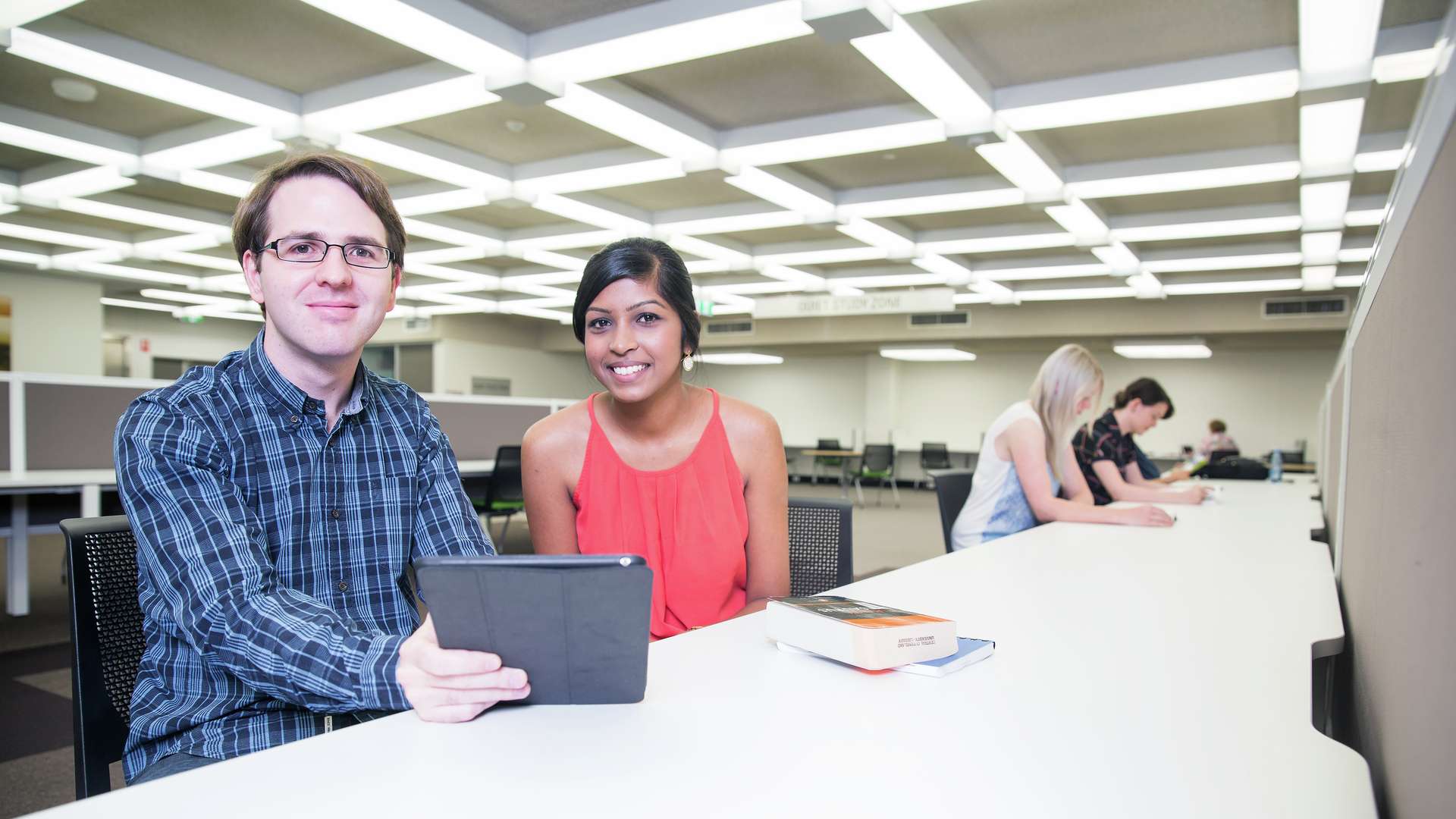 Two students look at a tablet and smile while other students study in the background.