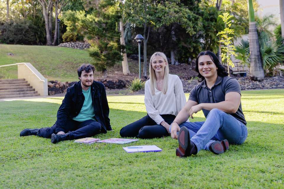 Three students sit outside in an open grassy area beside a colourful campus building