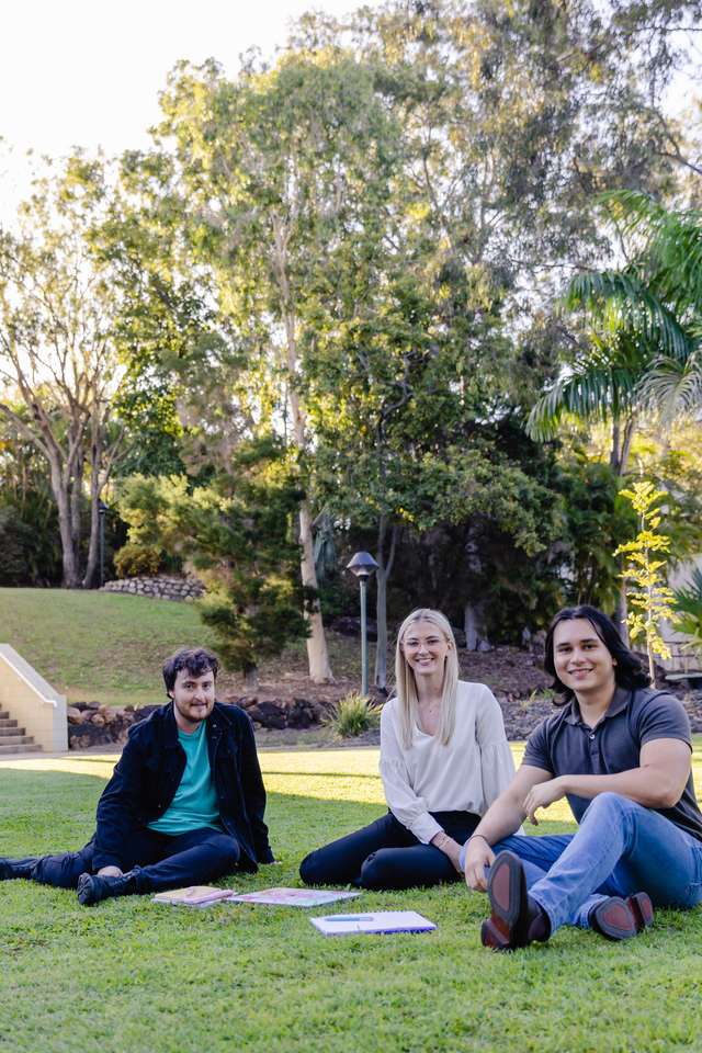 Three students sit outside in an open grassy area beside a colourful campus building