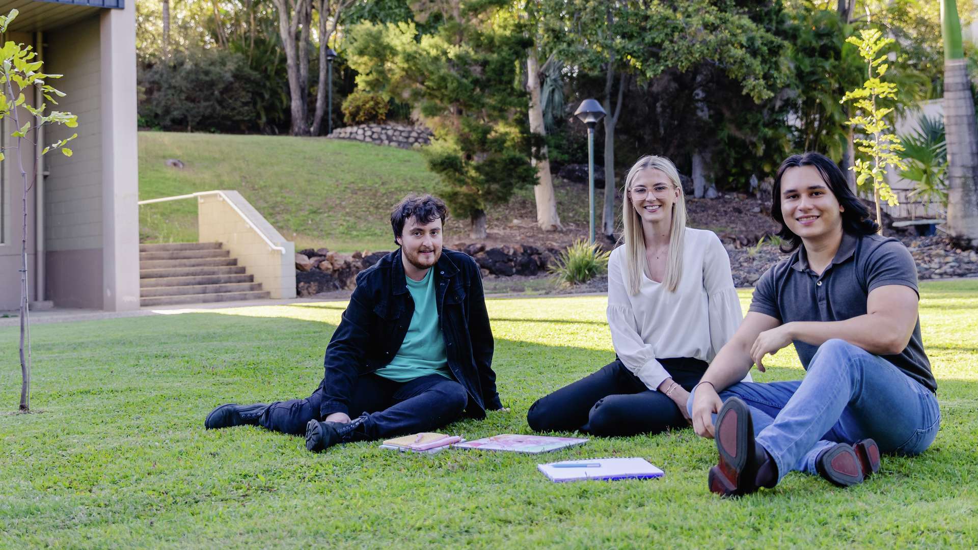 Three students sit outside in an open grassy area beside a colourful campus building