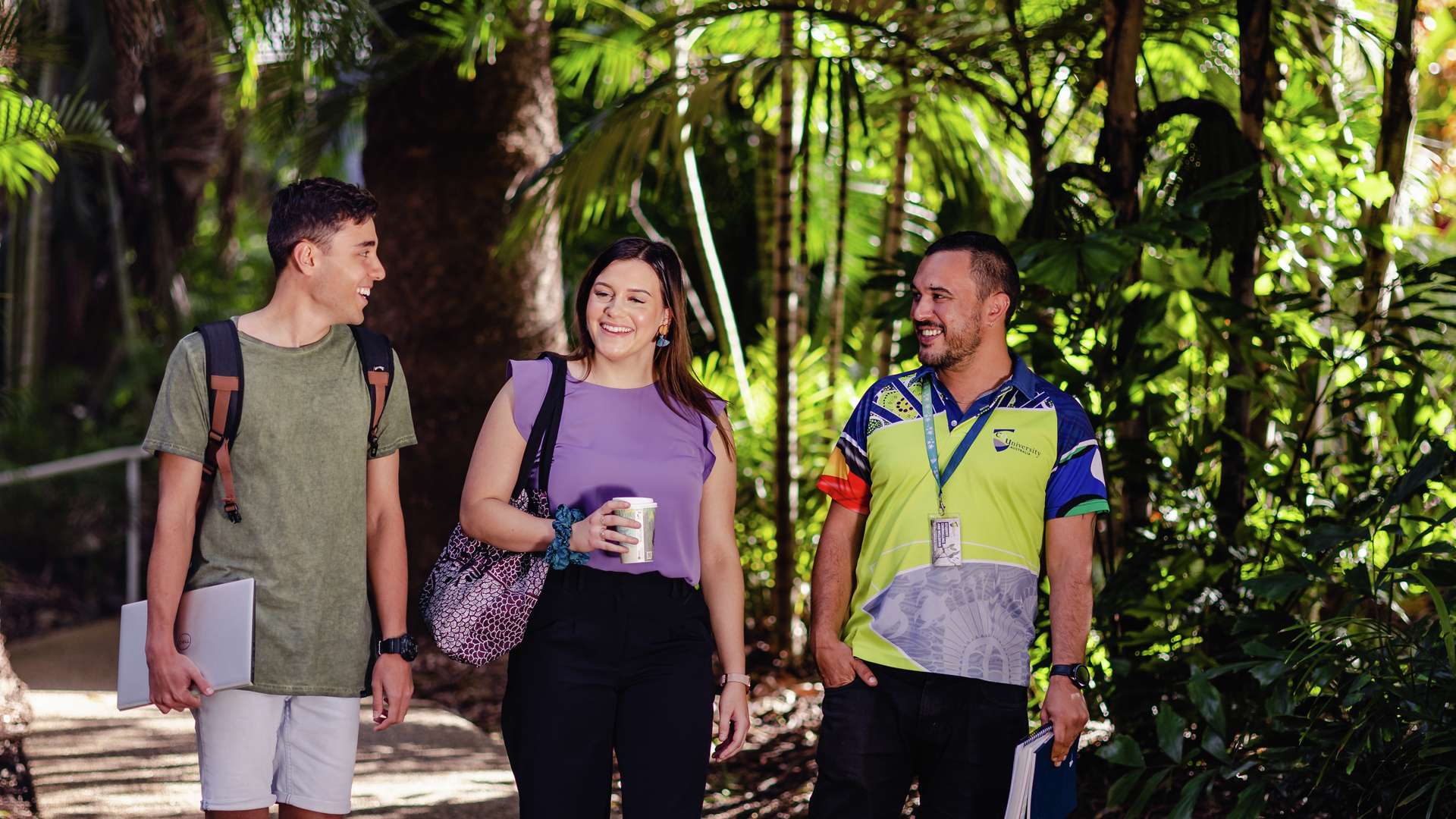 Three CQU students walking along path smiling, one holds a drink, two holding notepads