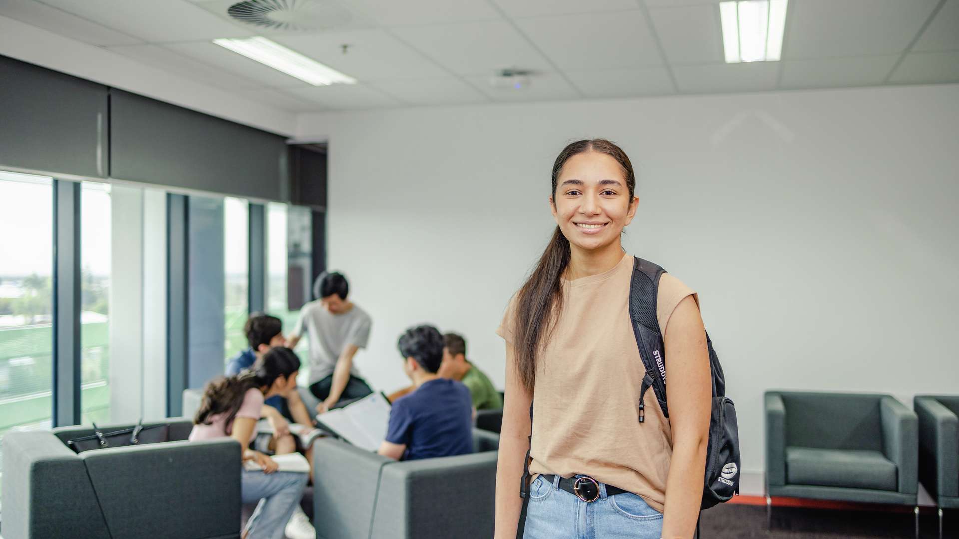 Student stands wearing a backpack in front of other students sitting studying in a group.