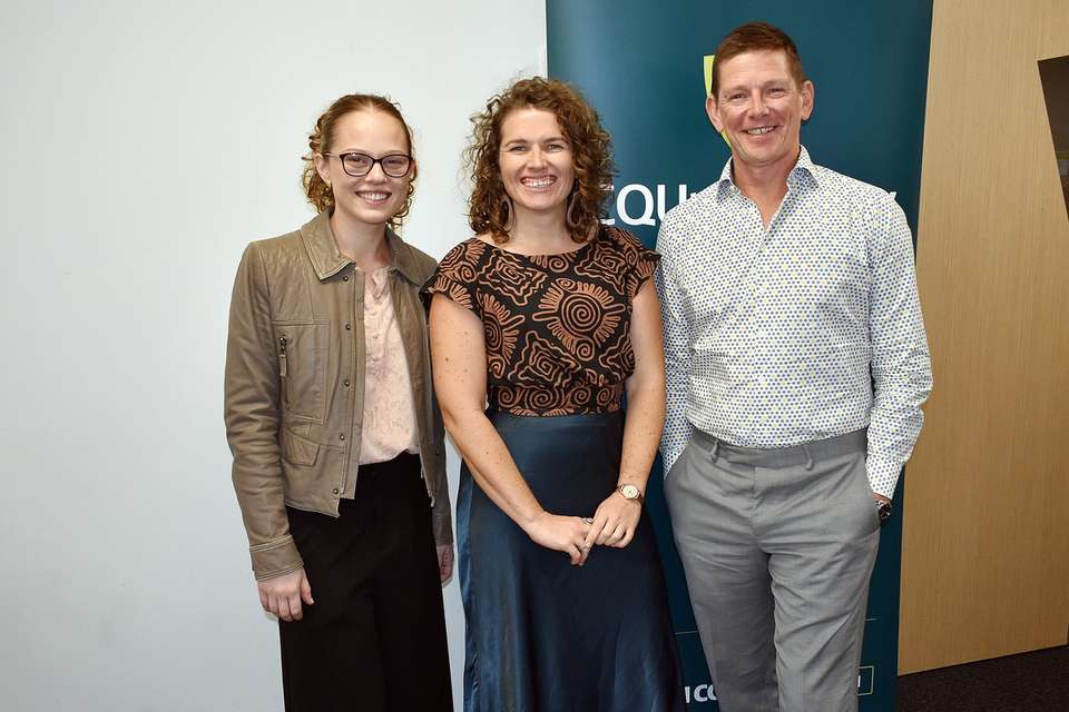 Three people standing together smiling in front of CQU sign.